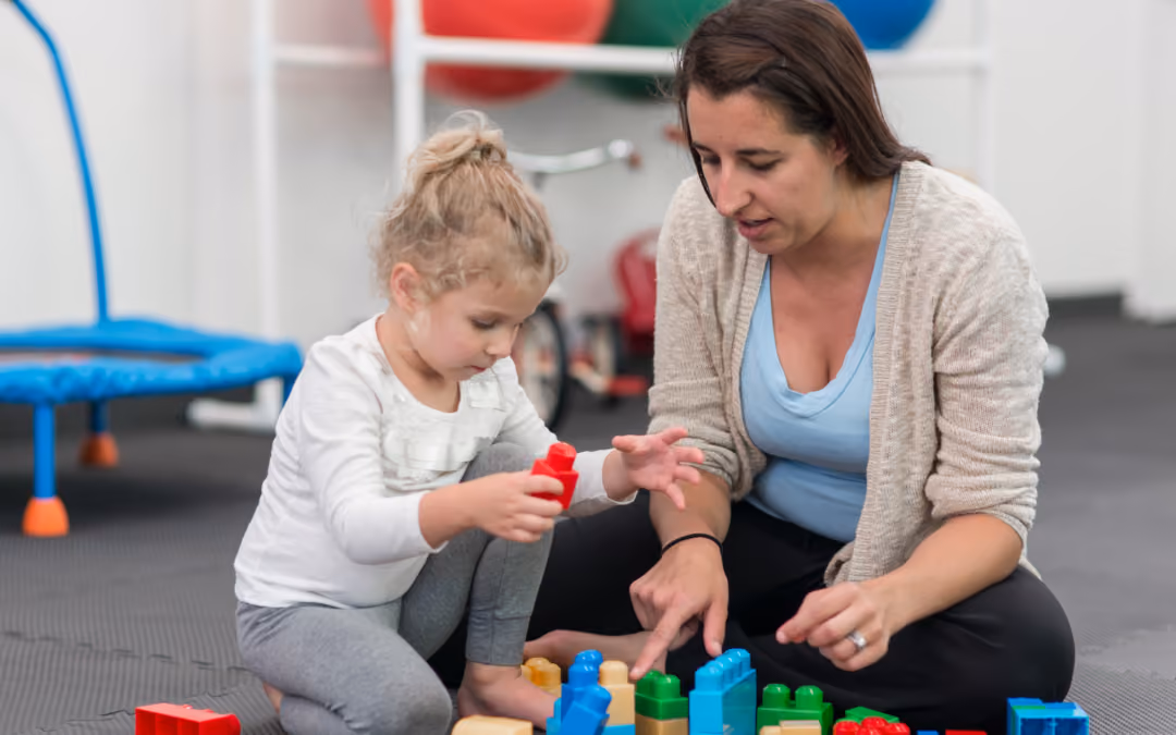 A clinician kneeling beside a toddler on a padded clinic floor, pointing at the colored blocks they are building with.