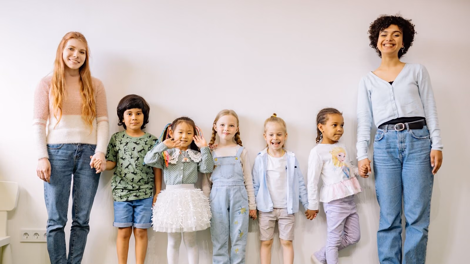 Two adults and six children standing in a line against a white wall on green turf, everyone smiling.
