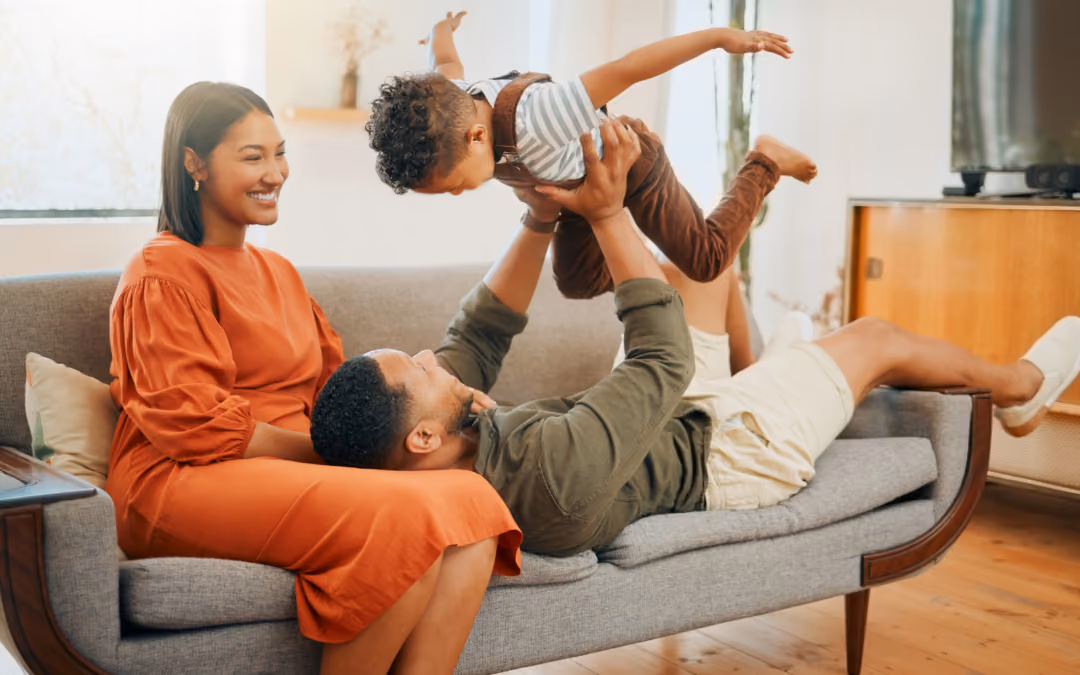 A family on their living-room sofa, one parent lifting a toddler overhead while the other sits alongside them, laughing.