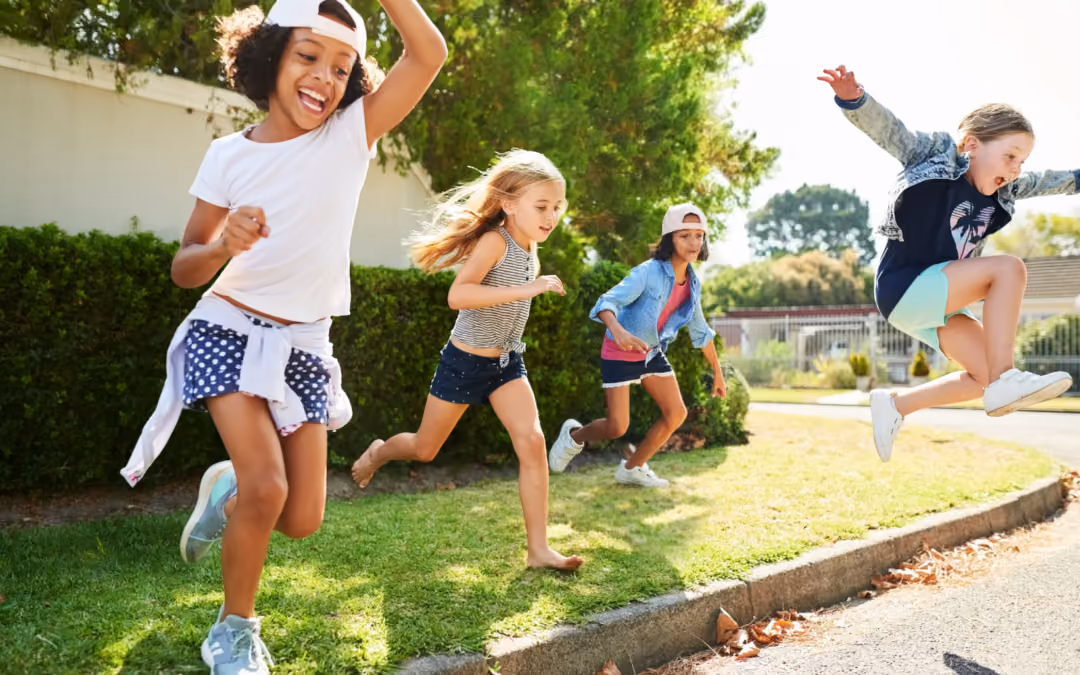 Four children running and jumping along a grass verge on a sunny suburban street.