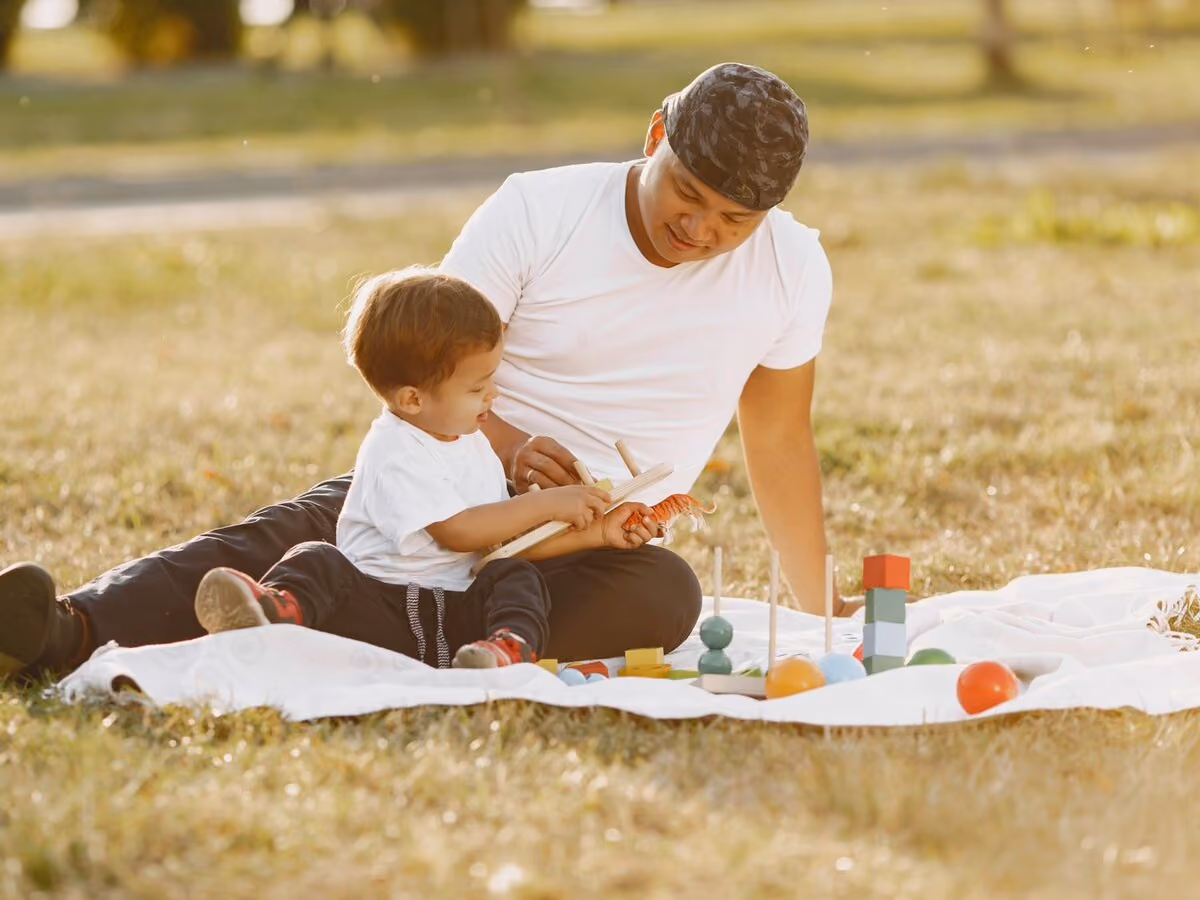 An adult sitting on a picnic blanket outdoors, working through a wooden toy with a toddler.