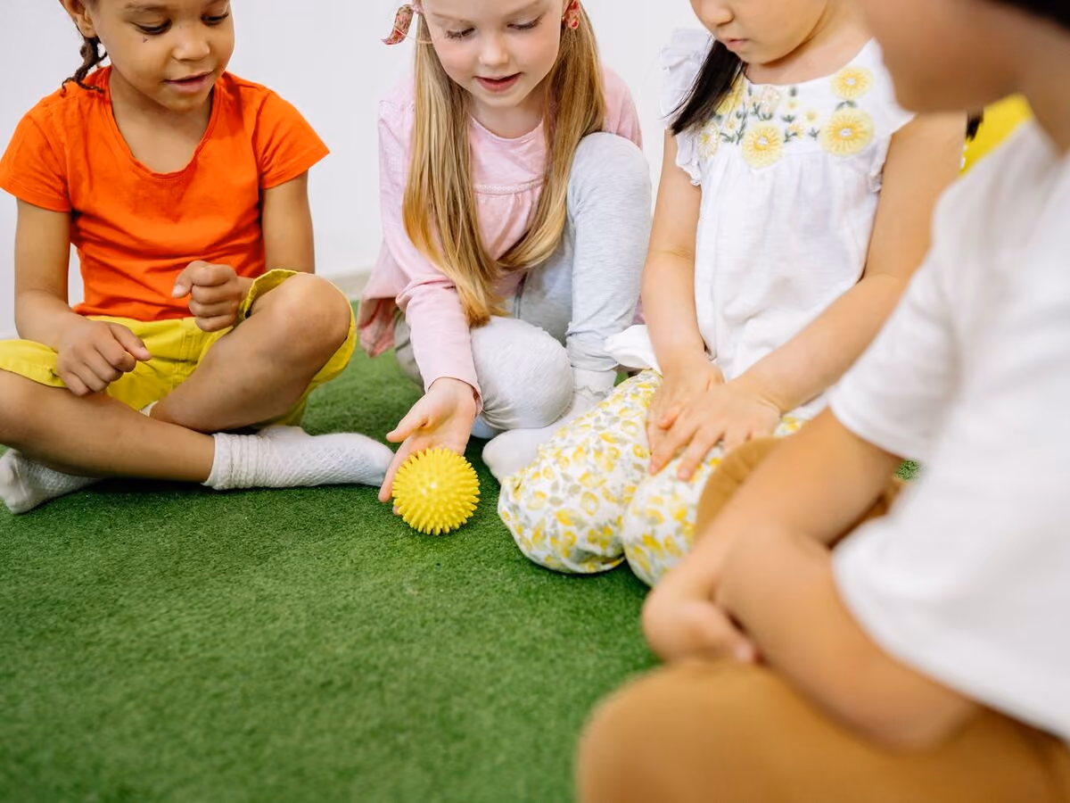 Four children sitting together on bright green matting, passing an orange spiky sensory ball.