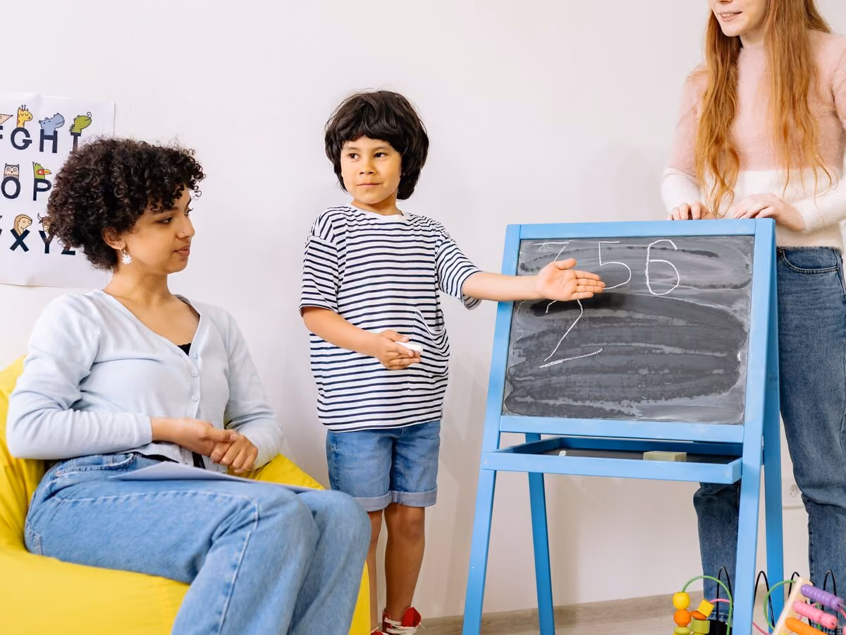 A clinician sitting on a yellow beanbag while a boy presents at a blue easel and a colleague looks on.