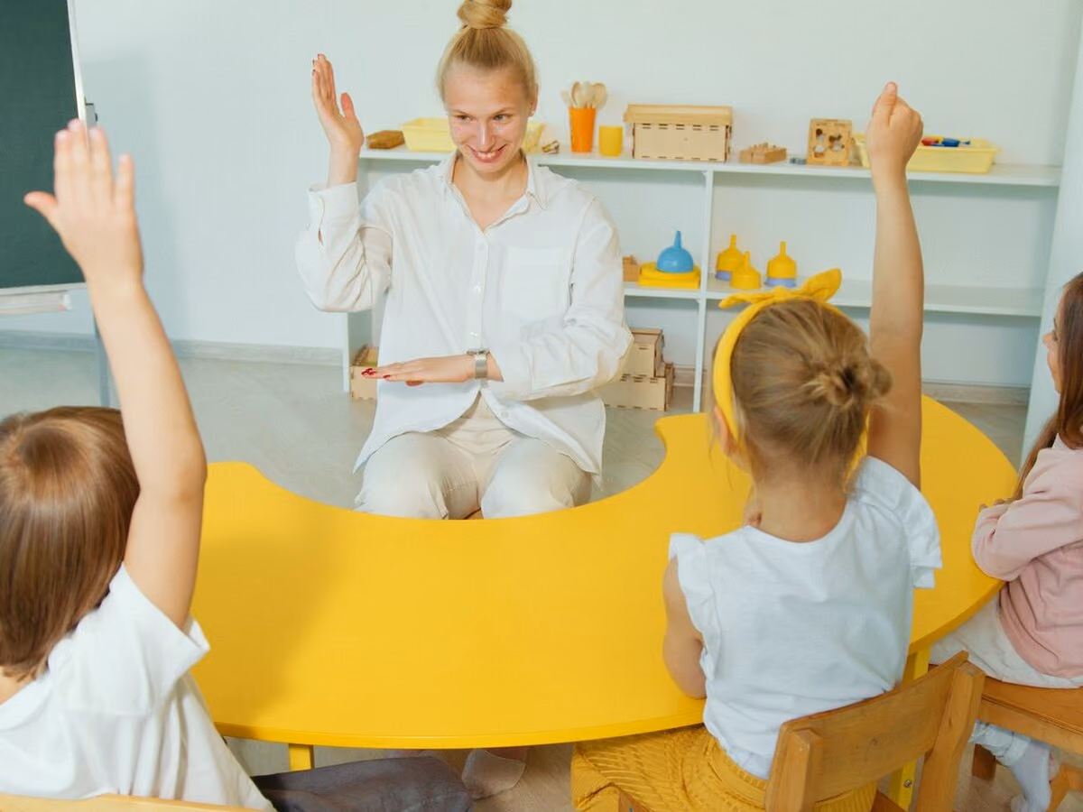 A teacher smiling at a bright yellow crescent table as three children raise their hands.