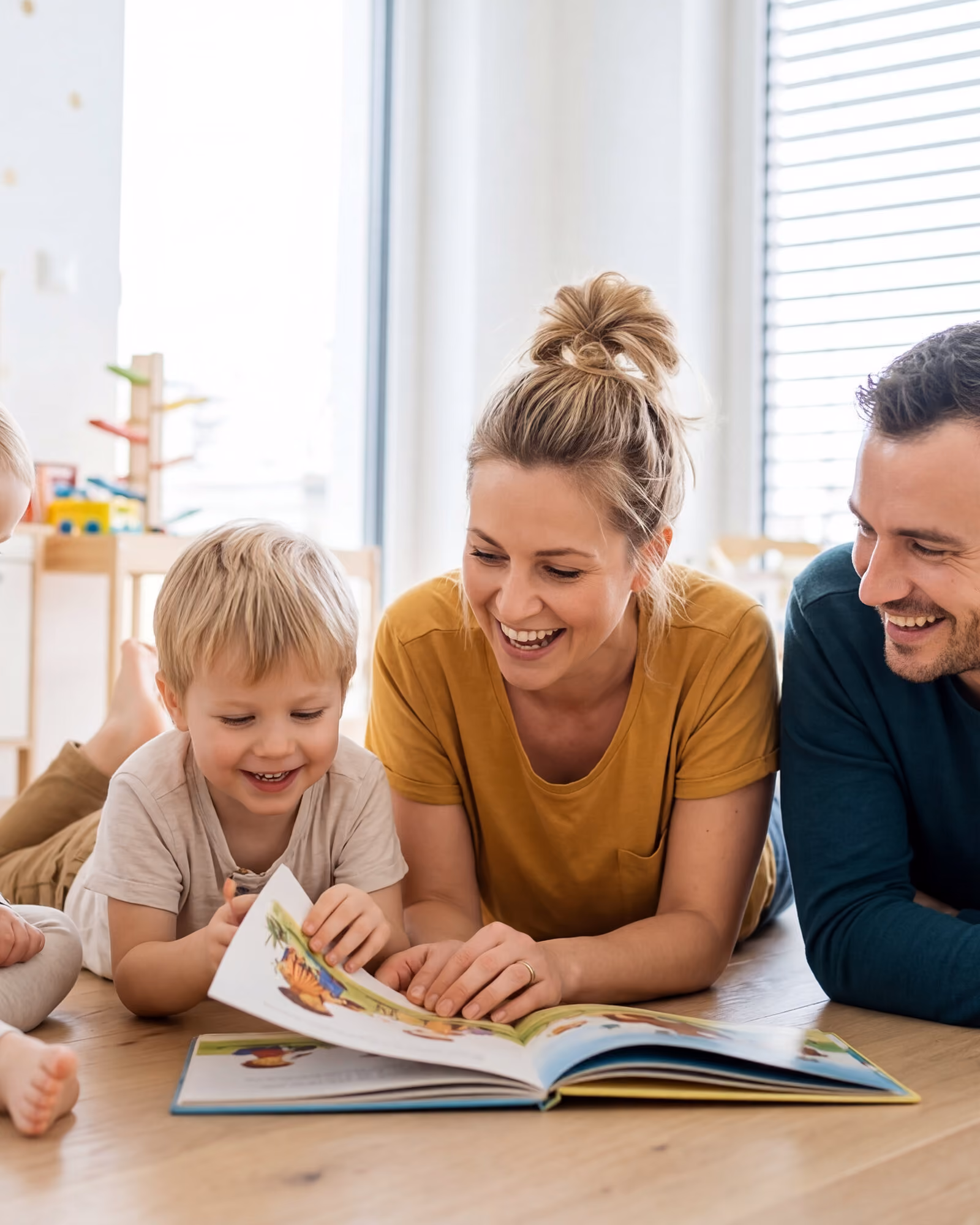 Two young children lying on a wooden floor over an open picture book, laughing, while their parents lean in over the page beside them.