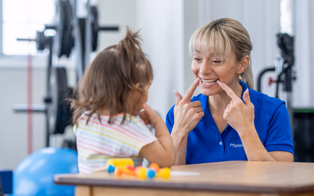 A therapist and a small child face to face at a table, the therapist framing her smile with both fingers as the child copies her.