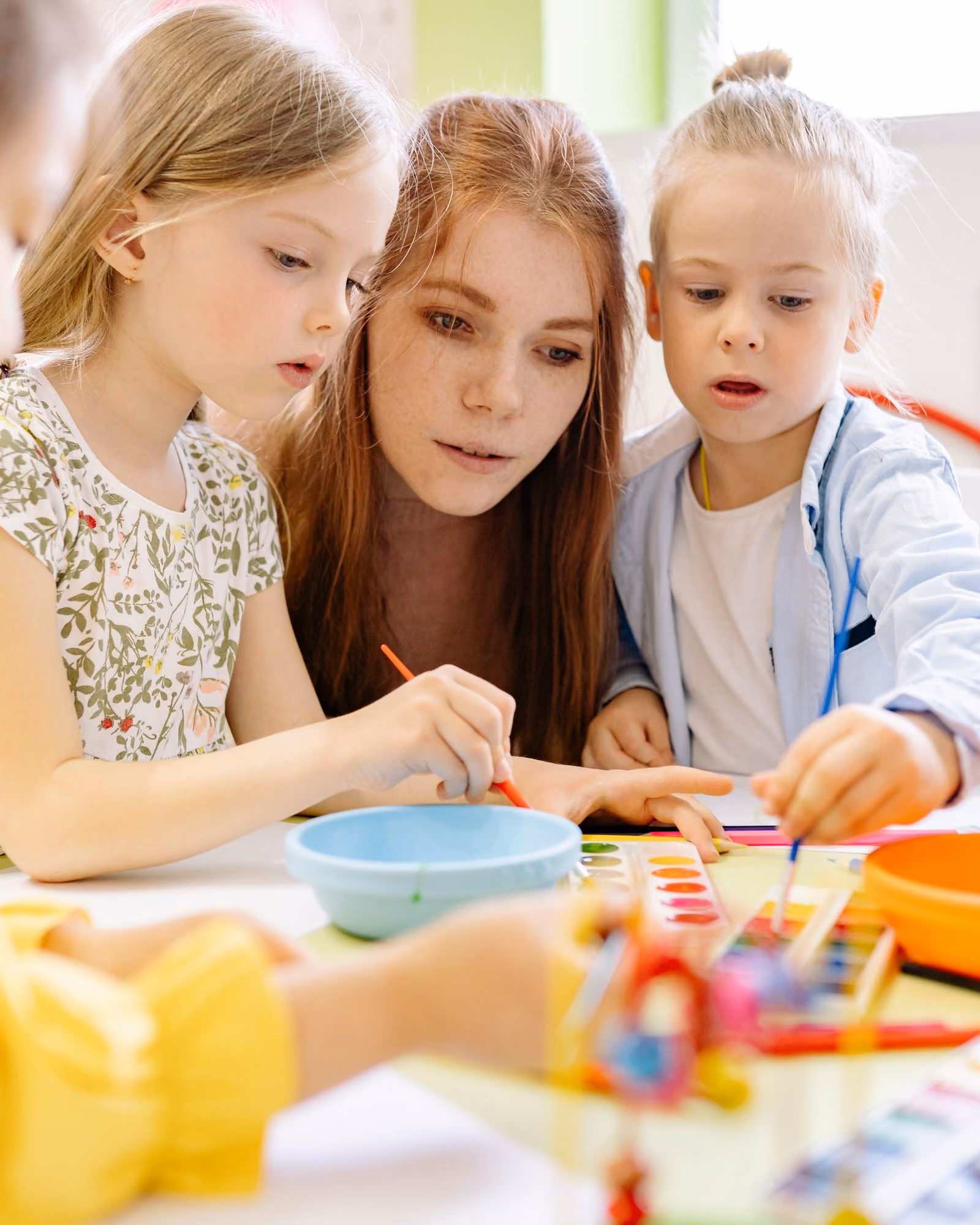 A therapist leaning in between two young girls at a bright craft table, painting together with watercolor palettes in front of them.