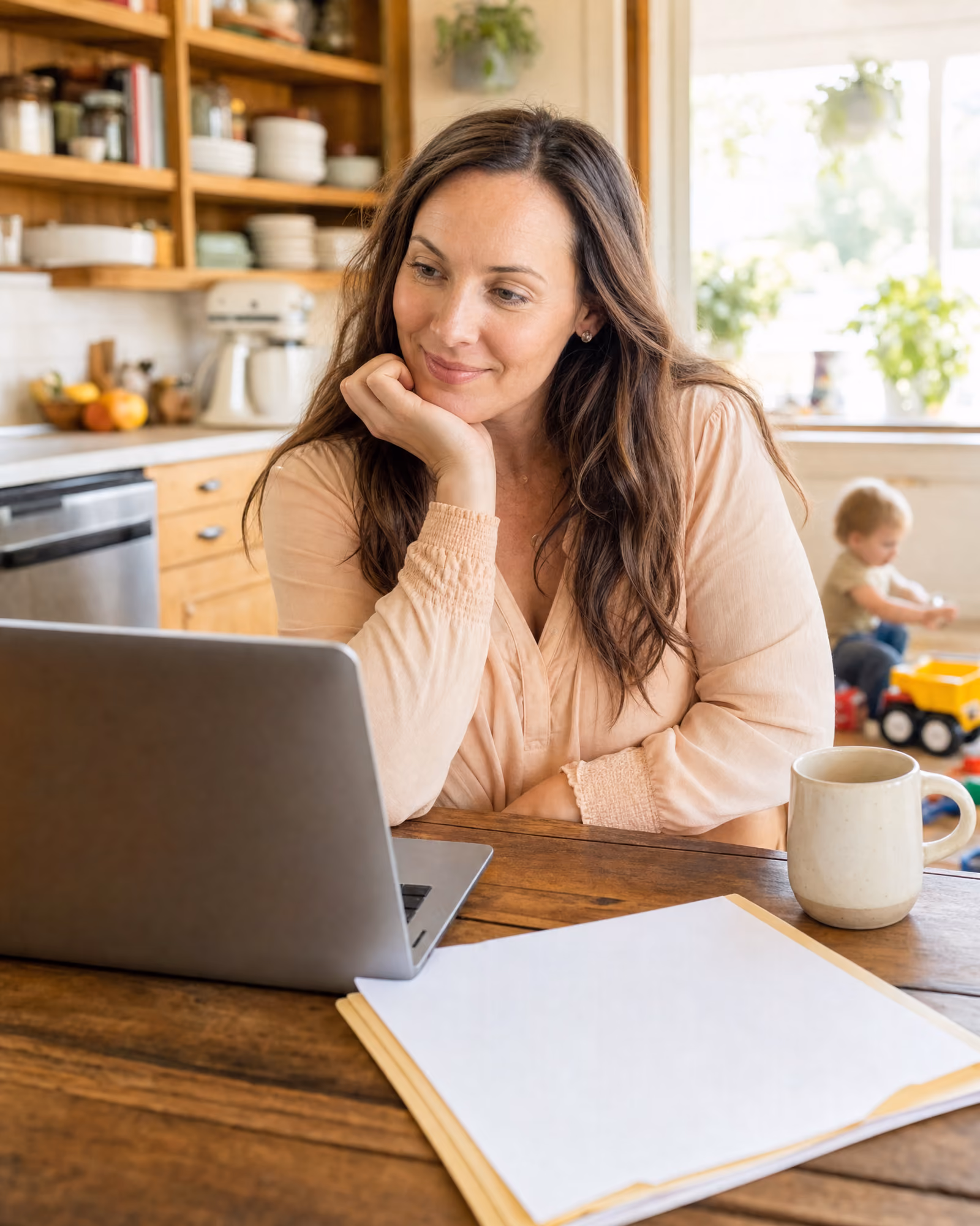 A parent sitting at a kitchen table with a laptop open and a folder of documents, working through paperwork in morning light.