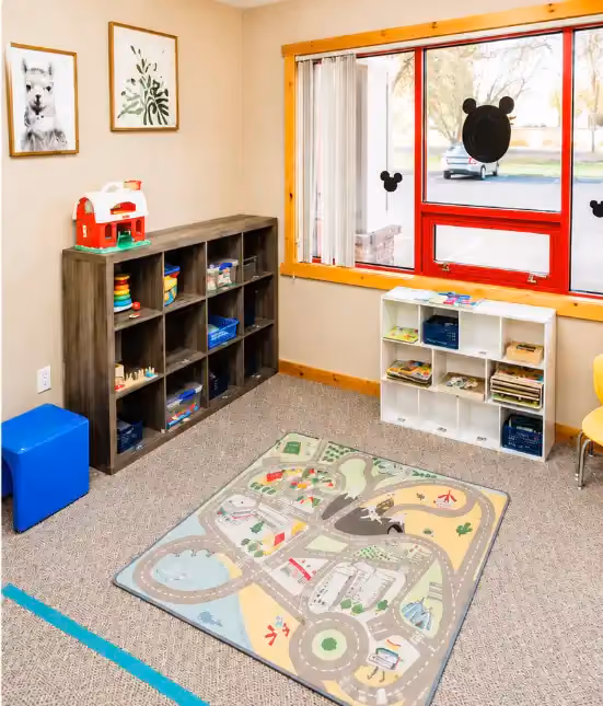 Toy shelves, a road-map play rug, and a red-framed window in the Idaho Falls clinic play area.