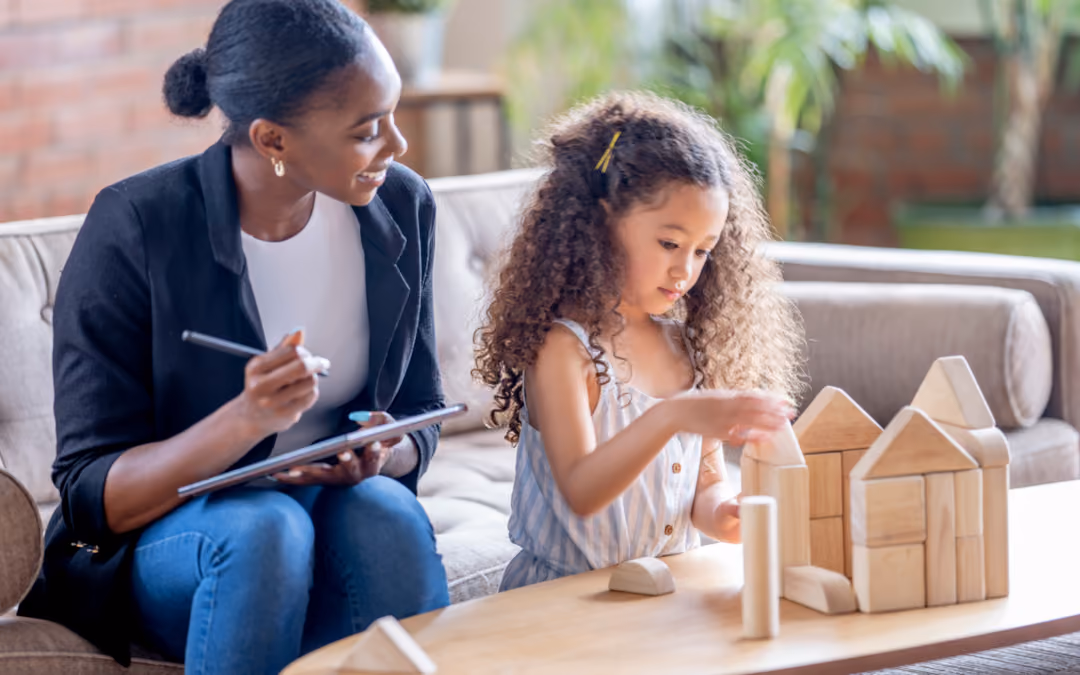 A clinician with a tablet watching a young girl build a house out of wooden blocks on a living-room table.