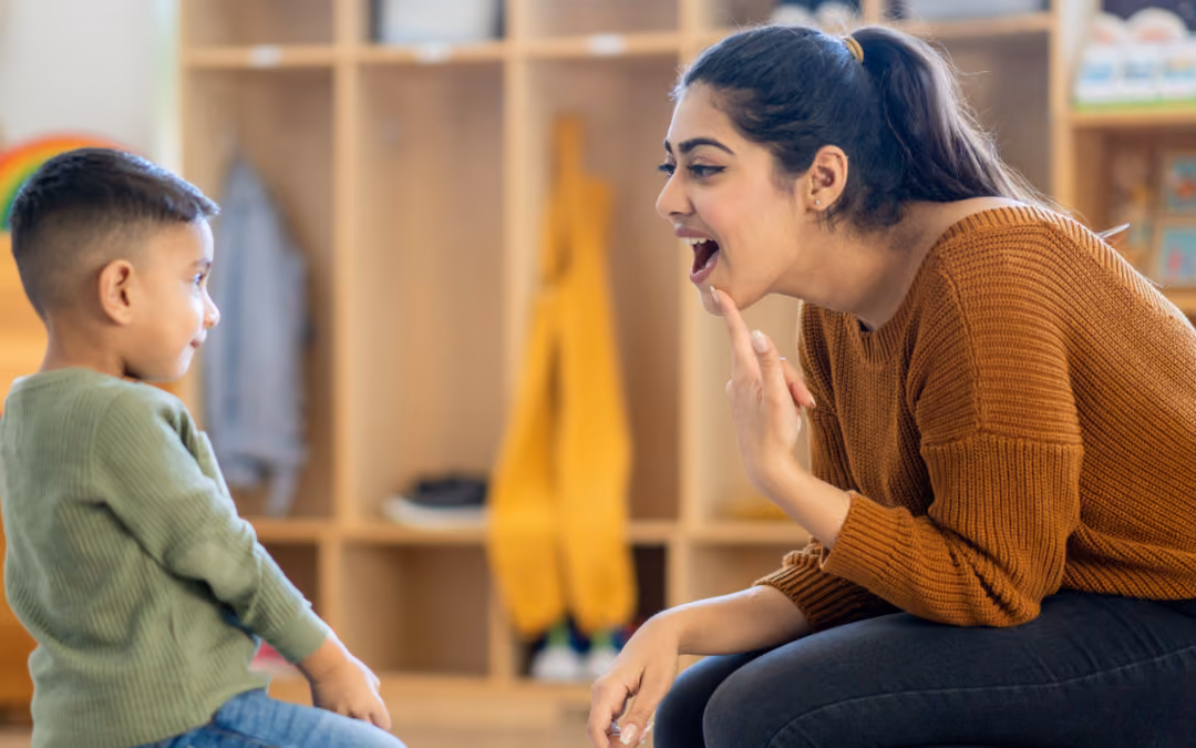 A therapist crouched to a toddler's eye level in a nursery, pointing at her own mouth to model the shape of a sound.