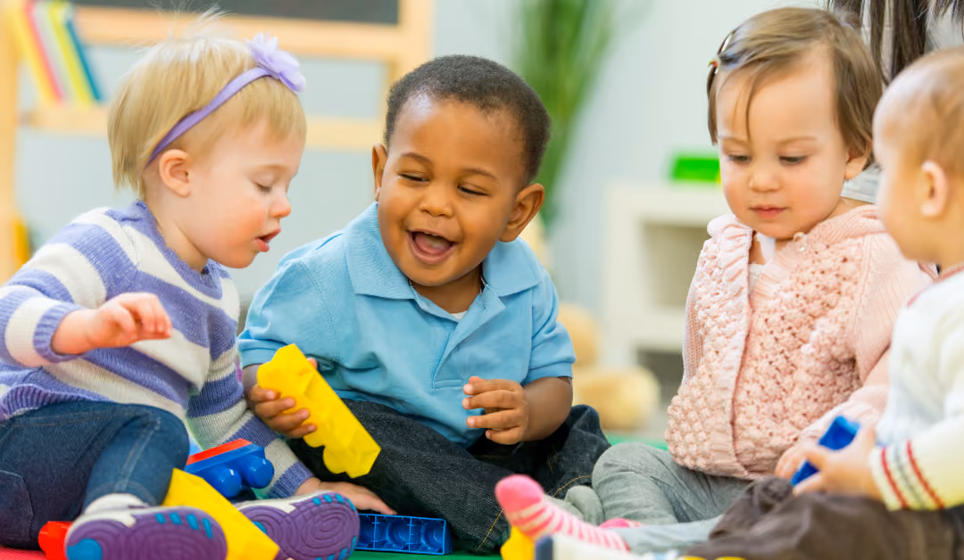 Four toddlers sitting together on a playroom floor, sharing a pile of plastic building blocks.