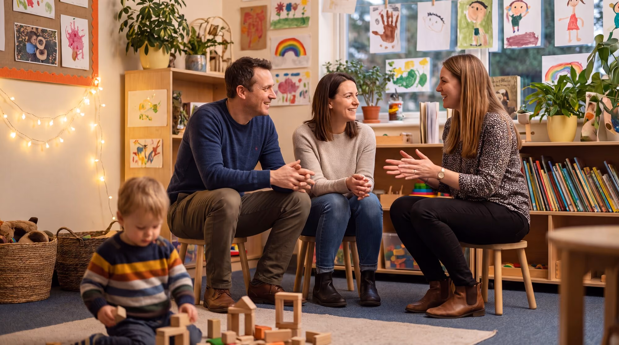 Two parents sitting with a Peak clinician in a bright playroom hung with children's paintings, while their toddler plays with wooden blocks on the rug.