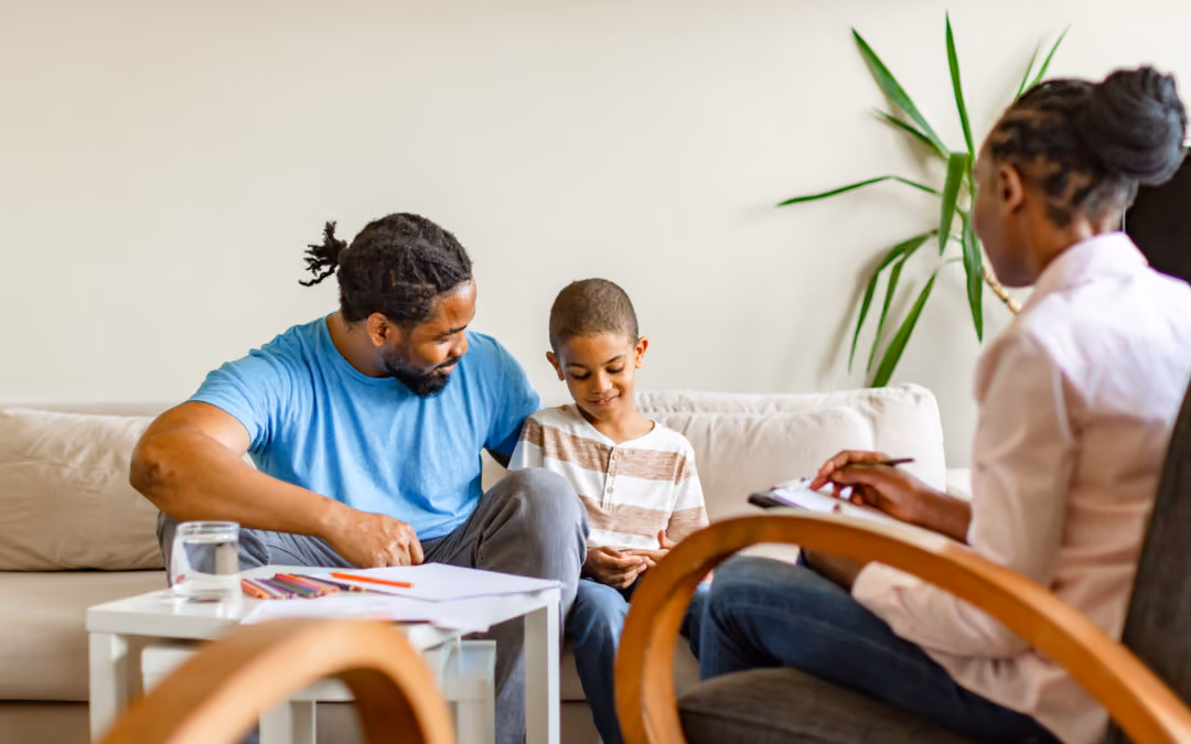 A father and his son drawing together on a sofa at home while a clinician sits opposite them, taking notes.
