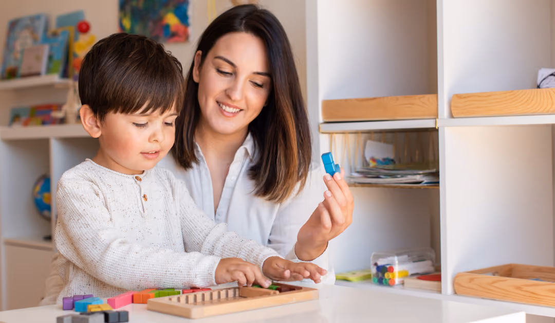 A boy fitting colored pegs into a wooden board at a classroom table while a teacher sits beside him and guides the next piece.