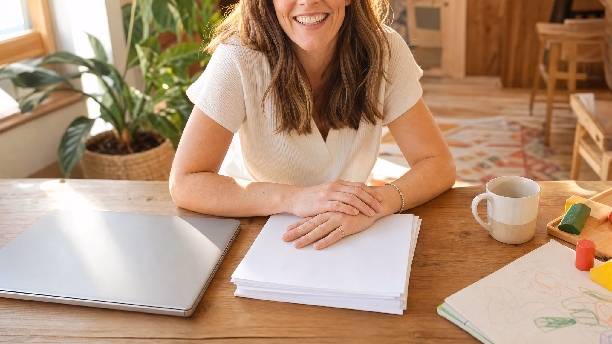A close view of hands resting on a stack of documents beside a laptop and a mug on a kitchen table.
