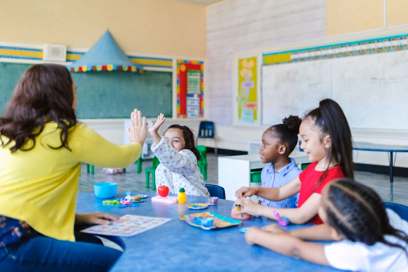 A teacher raising a hand to a child across a classroom table while other children work on an activity together.