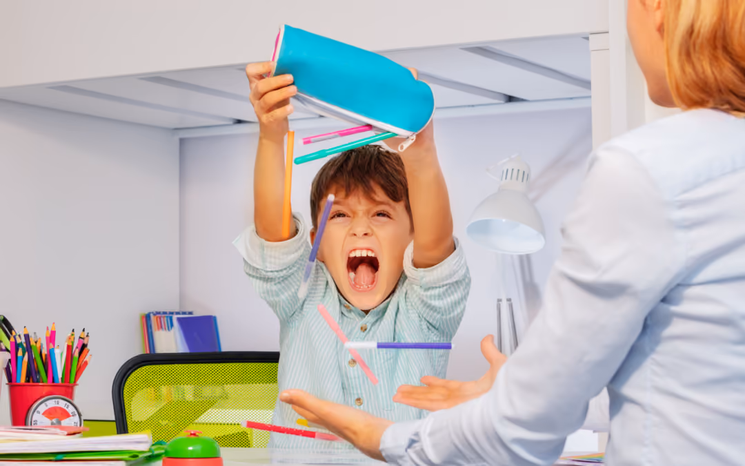 A boy at a desk tipping a pencil case over his head mid-shout, pens scattering, while an adult opposite him reaches out to steady him.