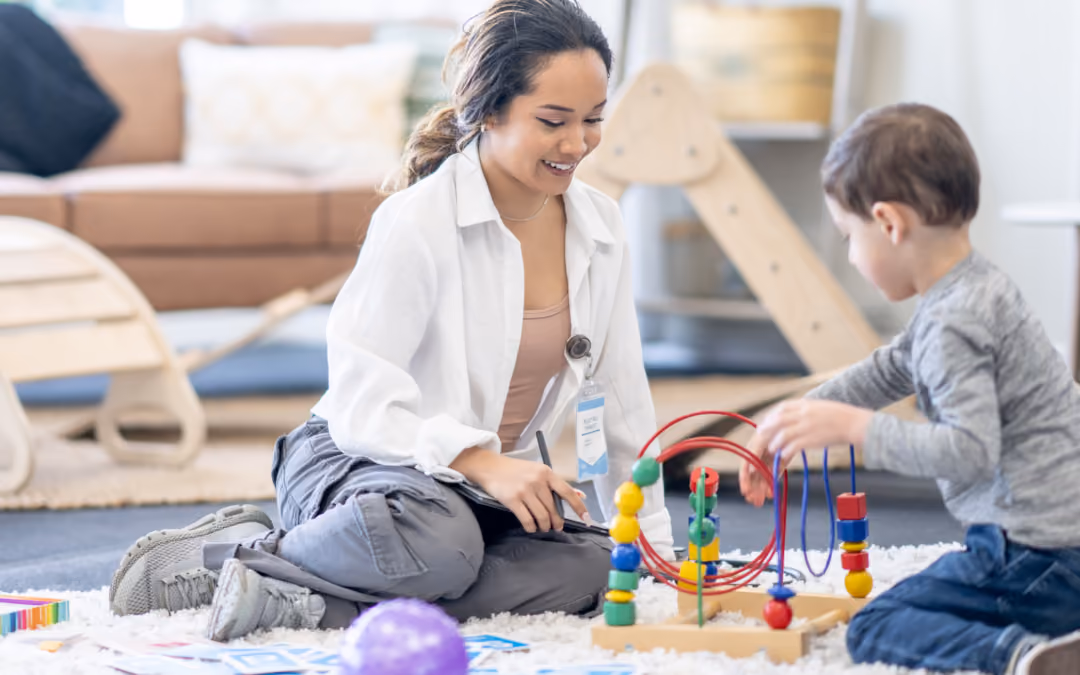 A therapist sitting on a living-room rug beside a boy playing with a bead maze, her notes and picture cards spread out on the floor.