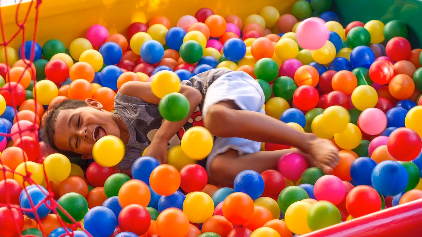 A child lying back laughing in a ball pit full of red, yellow, green and blue balls.