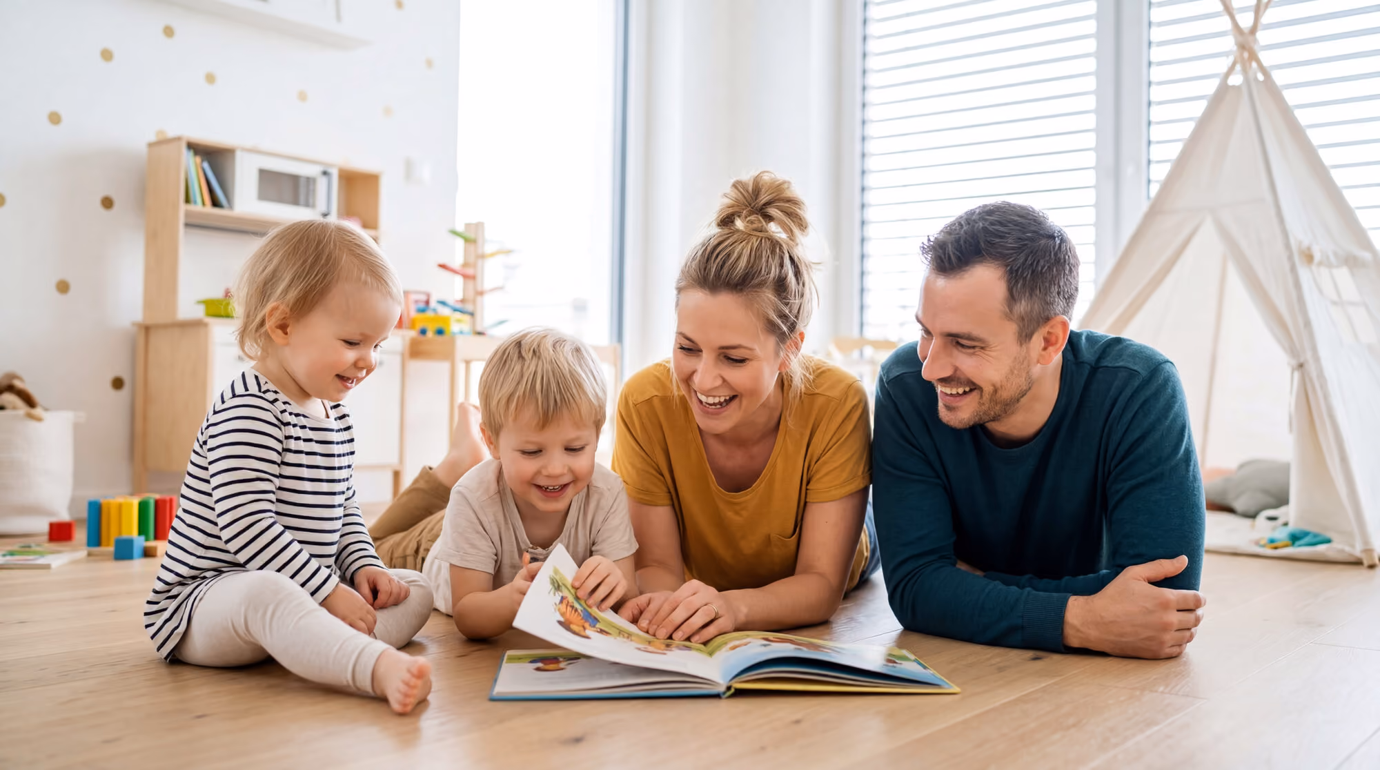 Two parents and their toddler and preschooler lying together on the floor of a bright, sunlit playroom, laughing over an open picture book.
