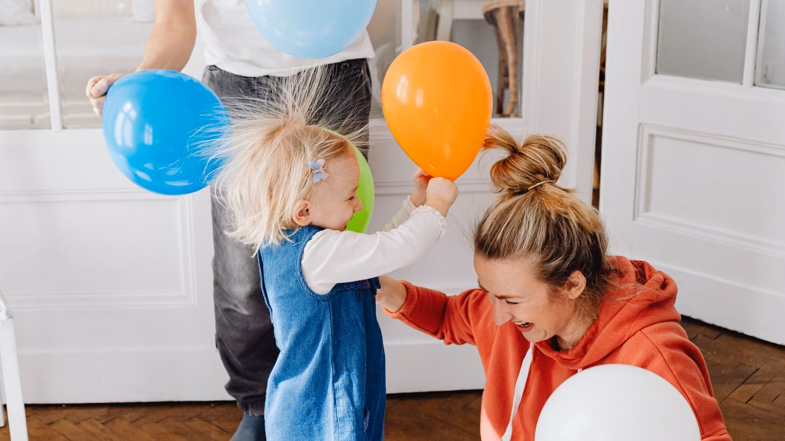A mother laughing with her toddler at home as they play with colorful balloons, the child's hair standing on end with static.