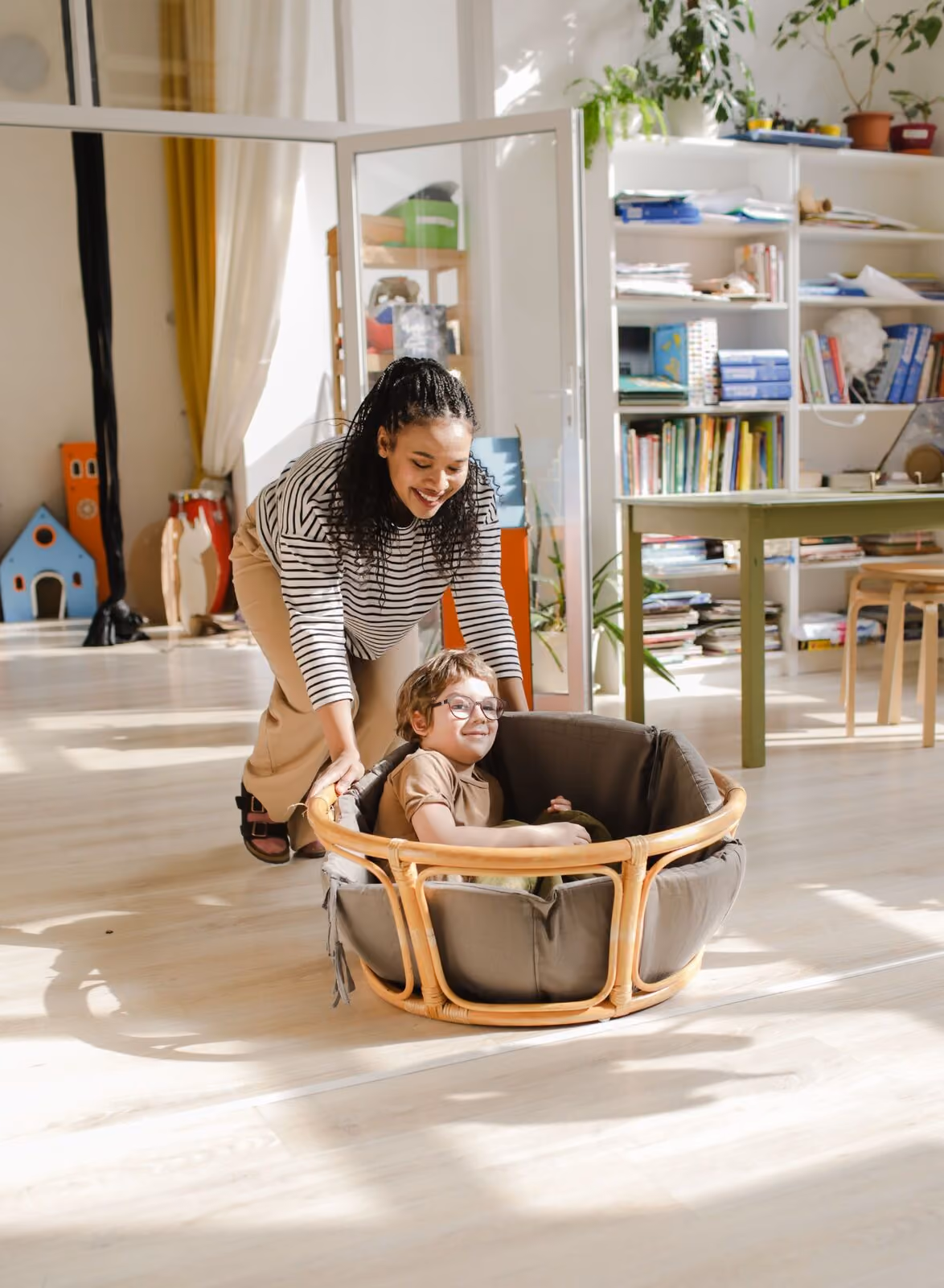 An adult pushing a laughing child along the floor in a round basket chair, in a bright playroom full of plants and toys.